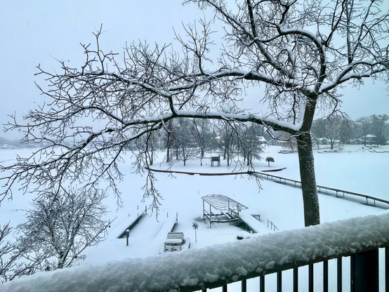 Photograph of a snow covered lake with snow, covered tree, branches across the top and a snow covered balcony rail across the bottom of the image.