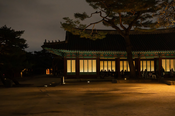 People rest in front of a Korean castle building at night. Windows are warmly lit.