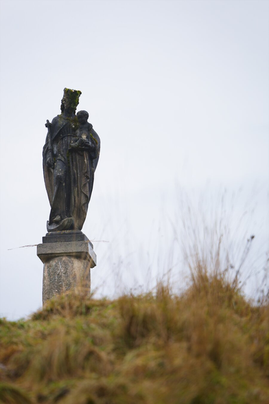 Stone statue of Mary (Christian figure) with Jesus, on a pillar on top of a grassy hill. White cloudy sky.