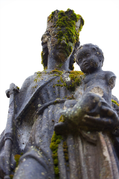 Closeup of a statue depicting Mary and Jesus, partly covered in moss. White cloudy sky.