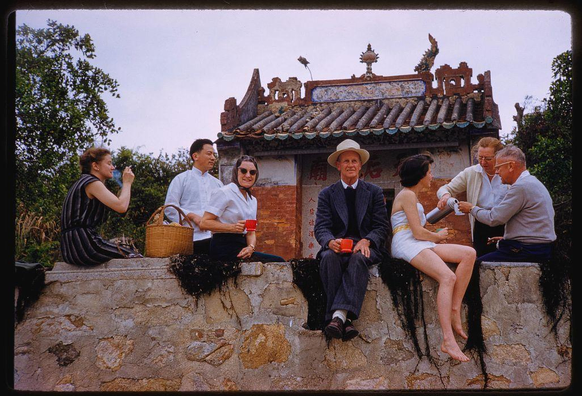 six people sitting on a brick wall in front of an asian building with tiled roof and decorative elements. two men hold red cups, another has a white cup, and one woman holds a basket next to her; the remaining three are holding teapots or serving containers. they appear engaged in conversation while enjoying tea at what seems like a picnic spot outside historical chinese architecture