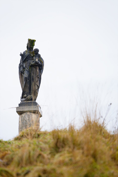Stone statue of Mary (Christian figure) with Jesus, on a pillar on top of a grassy hill. White cloudy sky.
