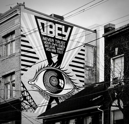 Mural on the side of a brick building. It is a stylised eye with diagonal lines radiating top and bottom in a wedge in between these lines at the top it reads “OBEY never trust your own eyes believe what you are told”. Other buildings are visible in the photo indicating an urban setting. The style is reminiscent of war propaganda, effectively communicating a feeling of surveillance and control.