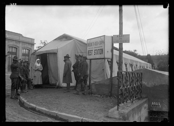 The image is a black and white photograph depicting an outdoor scene with several individuals gathered around temporary structures, which appear to be part of a field hospital or rest station. The tents are labeled "AMERICAN RED CROSS REST STATION." A signpost indicates directions towards other facilities such as the ABRI DE BOMBARDEMENT. In front of these makeshift shelters stands a group of people dressed in military and civilian attire from an earlier century, suggesting this photo could be historical.

The individuals around the tents seem to include soldiers wearing hats indicative of World War I-era uniforms, possibly American given the Red Cross reference. One woman is notably standing inside one of the tent structures; she wears white garments that resemble nurse's clothing, consistent with wartime medical personnel roles in such settings.

Additional details like a metal fence and pavement around the tents hint at an organized setup likely intended for supporting military operations or providing aid during war times. The attire and setting strongly suggest this image relates to World War I activities of American Red Cross workers near combat lines.
