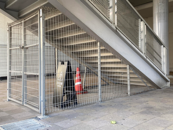 A metal fence surrounds a staircase in a building. A caution sign is positioned nearby, along with a traffic cone. The floor is tiled, and there are some fallen leaves.
