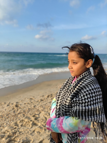 A young girl (Aylan) with long dark hair, wearing a black and white checkered scarf (keffieh) and a tie-dye jacket, stands on a sandy beach looking out at the ocean on a sunny day. The sky is blue with scattered clouds. The date and time are visible in the bottom right corner.