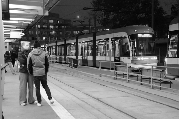 The outdoor platform of the station. Extending from the left background to the right midground are two streetcars in the far lane, brightly lit from inside. At left in the foreground, two men stand talking under the fluorescent lights of the platform.