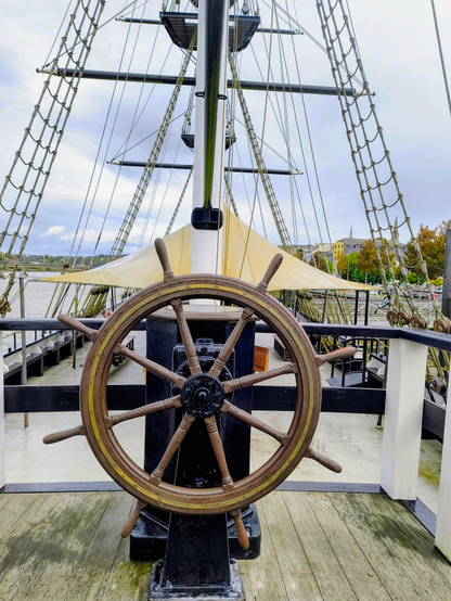 Die Mitte des Bildes füllt ein Steuerrad aus Holz aus, direkt dahinter ist ein weißer Mast zu sehen. Über all sieht man weitere Tagelage.