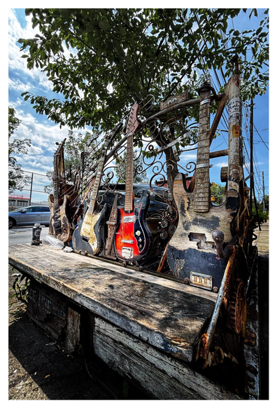 Photo of a wooden bench on a city sidewalk, with a backrest made of four dilapidated electric guitars stuck to some metal railing with swirling patterns. Three of the guitars are stringless and have scratched-up brown and black bodies; one guitar, second from right, has a still-glossy black-and-red body and has retained its strings. Various rusted metal odds and ends are also stuck to the railing, including a small rectangular metal plate with googly eyes. Behind the bench is a slender tree with green leaves. In the background are some parked cars, power poles, and a blue sky with white clouds.