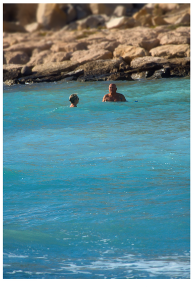 Hochformat: 2/3 des Bilds ist von bewegtem türkisen Meer ausgefüllt, dahinter unschärfer braungelbe Felsen. Unbeeindruckt vom Wellengang und Kälte stehen, fast schwimmen zwei Personen und unterhalten sich.