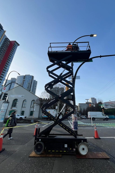 A worker is using a SkyJack lift to work on a streetlight. The image is taken on a city street with buildings and traffic visible in the background.