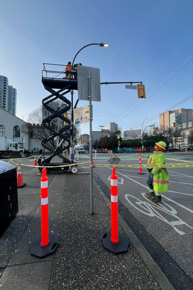 Workers are using a lift to work on a street light. Safety cones and caution tape are set up around the work area.