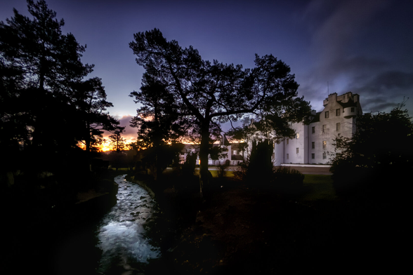 Ein historisches schottisches Schloss, umgeben von dunklen Kiefern, spiegelt sich im sanft fliessenden Bach im Vordergrund. Der Himmel zeigt die Farben des Sonnenaufgangs, von leuchtendem Orange bis zu tiefem Violett, während das Schloss dezent beleuchtet ist.
