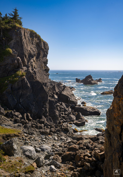 Color photo of a rocky beach in a gap between steep cliffs with the Pacific Ocean beyond under a clear blue sky. 