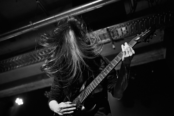 The image is a black and white photograph capturing a musician intensely playing an electric guitar during what appears to be a live performance. The central figure is seen from the chest up, with their head obscured by a mass of long, dark hair that is dramatically flying upwards and around, suggesting vigorous movement like headbanging.

The musician's hands are actively engaged with the guitar; one hand is on the fretboard, while the other is positioned near the body of the instrument. The guitar itself is dark, and the headstock, visible on the right, has "Harley Benton" written on it.

The background is dark and out of focus, with some metallic structures or stage rigging visible at the top. A bright, blurred light source is present in the bottom left corner, indicating stage lighting and adding to the dynamic atmosphere. The black and white treatment enhances the dramatic and energetic mood of the scene. (Aria)