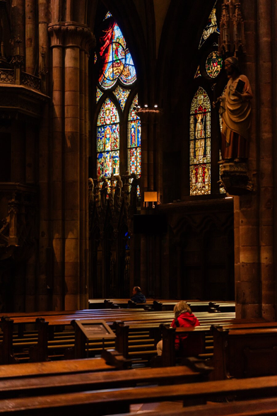 Interior of a cathedral featuring tall stained glass windows with intricate colorful designs on a dimly lit wall. Wooden pews occupy the foreground, with two individuals seated; one wearing a red jacket on the right, the other further back. Stone columns and a statue of a robed figure holding a book are visible on the right.