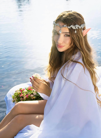 Elf-like young woman sitting by the water, wrapped in a sheer white fabric. She wears a silver jeweled headpiece with elf ears and holds a small flower in her hands. A bouquet of pink roses rests beside her on the white cloth, with sunlight reflecting on the lake in the background.