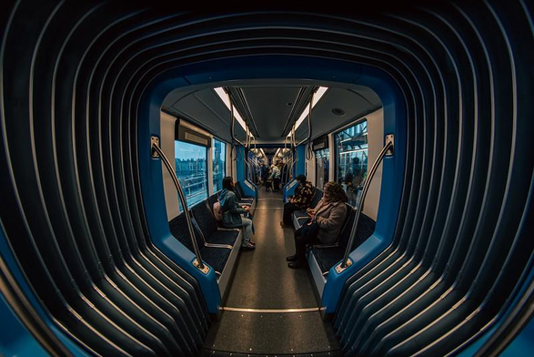 Fisheye shot looking down the length of a light rail train car. A short flexible section becomes a ribbed tunnel taking up half of the image, with the rest of the train car with people sitting and standing as they ride recedes into the far distance.