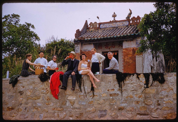 A group of people sitting on a wall next to an Asian-style building; they are all casually dressed, and some have picnic items with them. The photo was taken by Toni Frissell for Sports Illustrated in 1959 during sightseeing activities in Hong Kong. This particular image is stored among others in five boxes identified as F-J.