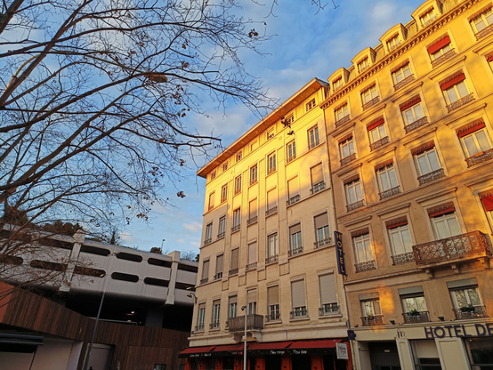 Une rangée d'immeubles du 19d siècle dont le haut est éclairé par le soleil levant. A leur gauche, une structure en béton percée d'ouvertures allongées (un parking) au-dessus d'un socle arrondi marron. On aperçoit sur le côté gauche de l'image les branches d'un arbre qui a perdu toutes ses feuilles.