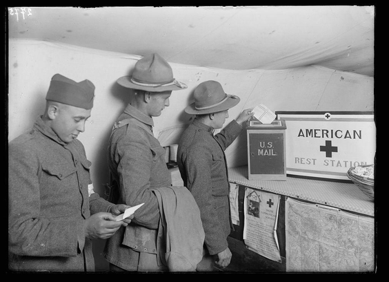 A black and white photograph depicting three uniformed soldiers engaging with a red cross rest station. One of the men is seen depositing an envelope into a mailbox marked "American U.S. Mail" while another looks on, holding papers in his hand. The third soldier appears to be focused intently at something out of frame.
The image captures a moment from military life during wartime or mobilization periods and highlights services provided by the American Red Cross for soldiers' mail needs.
