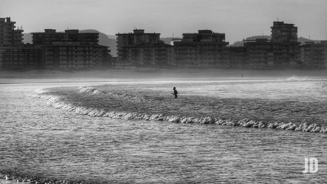 Fotografía en blanco y negro con una temática de playa y mar.
• Primer Plano: Se ve el mar con una ola rompiendo suavemente, creando una línea de espuma.
• Sujeto: Hay una persona, probablemente un pescador o alguien bañándose, de pie en el agua, a la altura de la cintura o el pecho, justo detrás de la ola que rompe.
• Fondo: Domina una larga fila de edificios de apartamentos o condominios de varios pisos, densamente construidos a lo largo de lo que parece ser la línea de costa o un paseo marítimo. Se aprecian montañas o colinas borrosas detrás de los edificios.
• Estilo: La foto tiene un ambiente brumoso o neblinoso, con un alto contraste, dándole un tono nostálgico o melancólico.