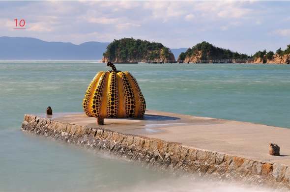 Photo couleur d'un paysage marin au Japon. Une digue s'avance dans la mer. Au bout de cette digue, une citrouille jaune à pois noirs. En arrière-plan des îlots rocheux couverts de végétation. Plus loin encore on apperçoit l'île principale du Japon. Le temps est beau mais le ciel est envahi de nuages.