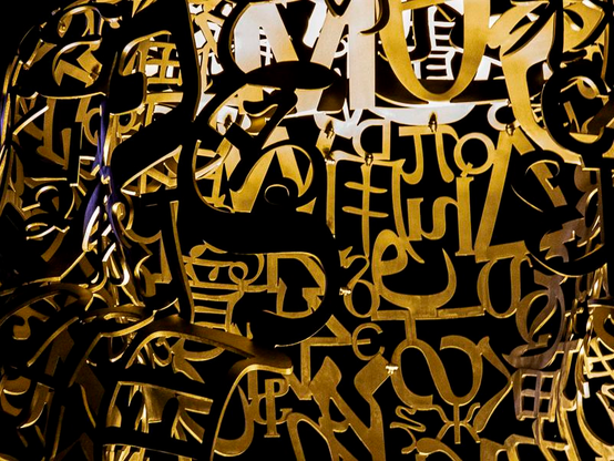 Une ohotographie au format paysage d'un détail d'une sculpture de Jaume Plensa.  Sur un fond noir un entrelacs à la fois serré et ajouré de lettres métalliques appartenant à différents alphabets et auxquelles la lumière artificielle donne un aspect doré. 