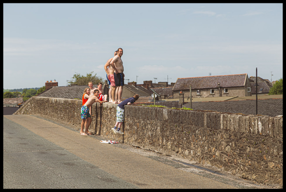 Photograph of a group of people in swimwear gathered on an old stone bridge in County Kilkenny, Ireland on a sunny summer day. Two shirtless men in swim shorts (one red/pink, one navy blue) stand on top of the stone parapet wall preparing to jump into the river below. A woman in an orange top and floral swim shorts leans over the wall alongside a child in a red and white striped top. Other children and young people in swimwear watch or prepare nearby, with one boy climbing the wall. Flip-flops and towels are scattered on the road surface. Traditional Irish stone buildings with slate and terracotta roofs are visible in the background under a blue sky with light clouds.