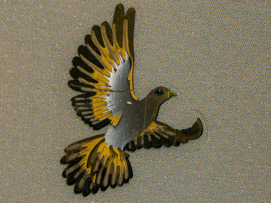 A silver dove on a mosaic background, with is wings spread. Due  to the lightning conditions, the feathers on the wings and tail are brown and golden. The visible eye is blue, and the beak has "VISA" written on it.