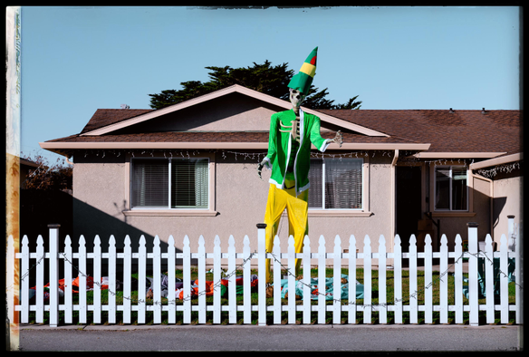 A photo of an ordinary, one story, stucco-coated, taupe-colored house with a brown shingled roof, white vinyl windows, and a white picket fence. Consistent with a late 1950s vibe, you can see Venetian blinds and drapes inside, in the windows. Outside, the house is decorated with Christmas lights: icicles from the roof eaves, mini LEDs how long the picket fence.

It's suburbia in a nutshell, if you were ignore the 15 foot tall skeleton standing in the front yard, dressed like an elf in green and yellow felt, standing over what appears to be the murdered bodies of Santa and some of his helpers.