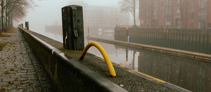 A bright yellow metal arch rests on a weathered stone wall next to a green mooring post along a misty canal. The foggy atmosphere obscures the brick buildings and bare trees in the background, while the water reflects the atmospheric scene.