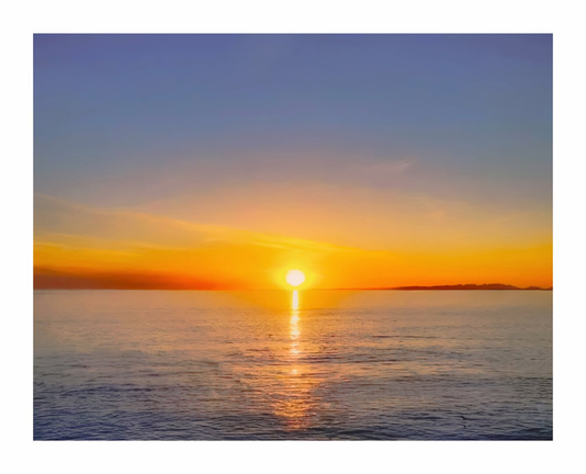 The is photo a scenic view of a sunset over the atlantic ocean (Galway Bay). The sun is positioned near the horizon with golden light that reflects off the water's surface. The sky transitions from a blue at the top to a warm orange near the horizon. A distant landmass (Galway) is visible on the right hand side of the image.