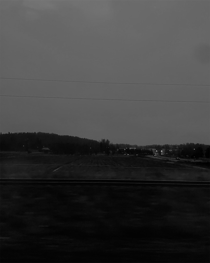 A dimly lit landscape at dusk, featuring distant hills, faint lights in the background, and a foreground of dark fields.