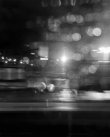 Abstract black and white image of blurred lights and raindrops on glass, creating a moody, out-of-focus cityscape at night.