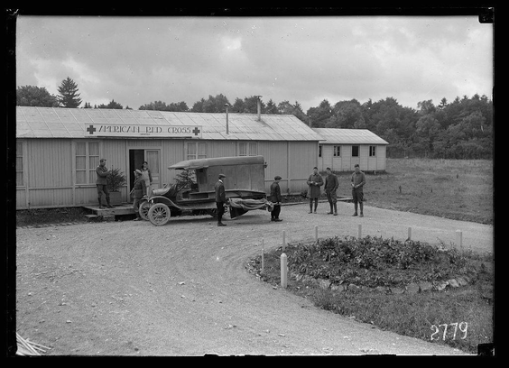 The image is a black and white photograph depicting an outdoor scene in front of two buildings labeled "AMERICAN RED CROSS." A vintage truck with what appears to be medical equipment or supplies on its bed is parked outside one building, with several men gathered around it. Some are carrying objects that could be related to the Red Cross's activities.

The environment looks rural with a grassy field and trees in the background under an overcast sky. In front of the truck, there is a circular garden patch containing plants or crops enclosed by two metal posts connected via chains. The ground appears unpaved, consisting mostly of dirt with scattered rocks.

Additional context for this image can be found at "Patient arrives at Hospital (ARC) Neufchateau," which suggests that this photograph might have been taken during wartime when the Red Cross provided medical care and support to those in need. It is indicated as part of a series or collection, possibly illustrating different aspects of humanitarian aid work associated with the American Red Cross during such events.

The image identification number "2779" could be used for cataloging purposes within an archive system.