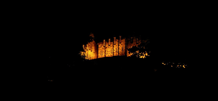 Night photograph of Edinburgh Castle, brightly illuminated in golden light, silhouetted against a completely dark sky. The stone walls and windows of the castle buildings are visible above the dark foliage of trees in the foreground.