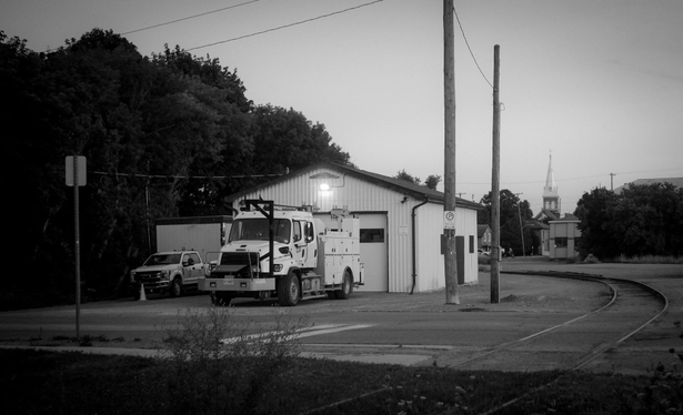 A large white work truck and a smaller white pickup truck stand facing the photographer in front of a building. A outdoor light shines on the building and large trees can be seen to the left and tall utility poles on the right, the ground is paved. Railway tracks can be seen curing behind the building as well as a church steeple in the distance. This is a black and white image. 