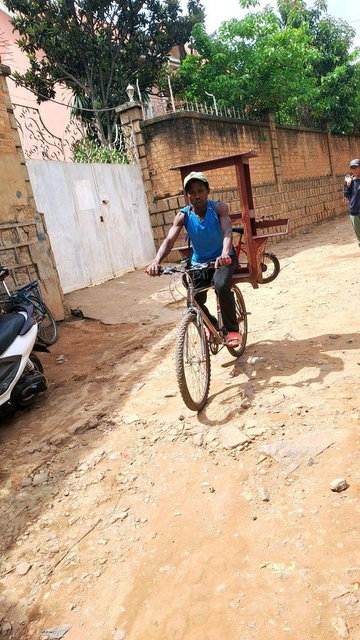 A man rides a bicycle on a dirt road in Antananarivo, Madagascar, with a tall, narrow wooden table unit strapped to his back. Two shelves near the bottom, gives the appearance of wings. Behind the bike, a child’s bicycle is also being hauled, showcasing the creativity and resourcefulness of transporting goods by bike.