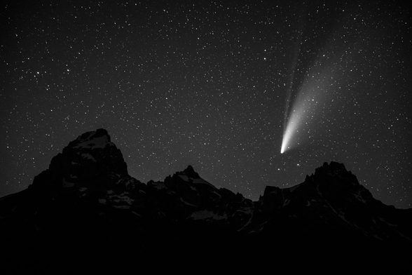 Comet NEOWISE setting behind Teewinot Mountain in Grand Teton National Park. Grand Teton, Mount Owen, and Teewinot Mountain are seen in silhouette under a very starry sky, with only patches of snow barely visible on the mountains.