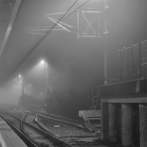 Low-contrast square photo of a platform and two intersecting tracks. The beams of light from two lanterns are faintly visible in the fog.