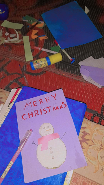 A handmade Christmas card with a watercolor snowman wearing a pink scarf and written "Merry Christmas" on top of a blue folder. Other crafting supplies, including markers, glue, and cut paper are scattered around.