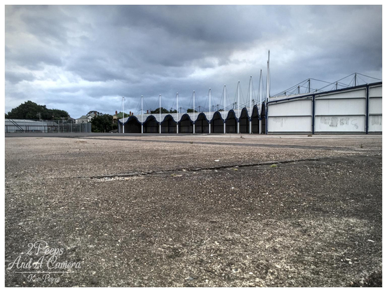 A low angle, wide shot of a large, desolate, cracked asphalt or concrete expanse leading toward the industrial sheds of a former rail yard under a heavily overcast, moody sky.

In the mid ground, a long, curved row of shed entrances with a distinctive, pointed, scalloped roofline provides a strong geometric focal point, flanked by a large white building on the right.