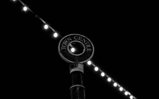 A black and white photo, with a string of lights in the background, with a round sign in the foreground. The sign is on the top of a post, circular, and has the text. "Town Centre" written within it. 