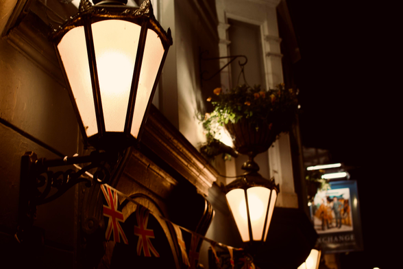 An image of the front of a Pub. It has classic gas lantern looking lamps out front, as well as bunting of the UK union flag.