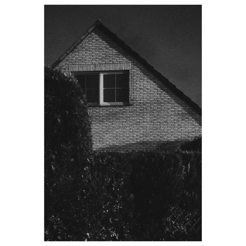 Schwarz-weiß Foto einer Nachtszene. Hinter einer hohen Hecke ragt die hell geklinkerte Giebelseite eines Wohnhauses vor dem dunklen Nachthimmel auf. Unter dem Dach sieht man ein Sprossenfenster sitzen 