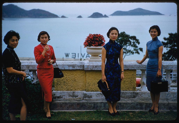 A color photograph shows four women posing in front of a stone railing overlooking the sea. The background features an expansive body of water with several small islands visible on the horizon, and there is a large potted plant with red flowers between two standing women dressed in blue dresses. 

The woman to the far left wears dark clothing with patterns resembling snakeskin or reptilian skin textures; she carries what appears to be binoculars around her neck and holds a black purse at waist level while smiling towards the camera.

Next is a woman clad in a bright red dress, paired with high heels, holding up one fist as if waving or gesturing. She accessorizes with pearl earrings and necklace set against an olive-green backdrop suggestive of foliage beyond the railing.

The third standing female to her right wears a striking blue floral-patterned outfit that features short sleeves, complementing it with dark stockings; she holds onto another black purse while looking directly at the camera.

Finally, on farthest right is one woman in a mid-length navy-blue dress and heels holding a matching briefcase-style bag. She exhibits a mild smile as well, her gaze directed toward someone off-camera's left side. The women appear to be standing on an outdoor terrace or promenade near steps leading down towards the waterfront.

The setting seems informal yet posed for photographs capturing their enjoyment of sightseeing in Hong Kong during 1959.