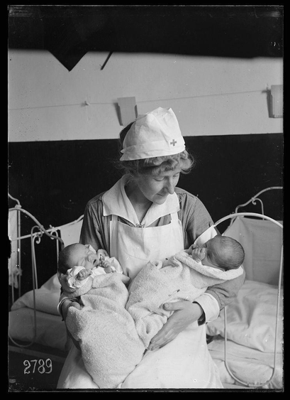 The image is a black and white photograph depicting an elderly nurse holding two newborn babies wrapped in blankets. She wears a nursing cap with a red cross emblem, indicating her role as a healthcare worker or midwife during the early 1900s, possibly around World War I based on historical context provided.

Details: The central figure is presumably named after Lewis Hine's work from June 1918 at Asile Caserne du Luxemborg. This setting suggests it may be part of a series documenting social welfare and maternity care during wartime or its aftermath. Additional background includes hospital beds, which support the context as medical facilities.

Historical Context: The image references World War I, indicated by information mentioning "Father was killed at the front 4 months ago." It portrays maternal care provided to newborns whose father has passed away in war, underscoring wartime impact on families and social welfare measures.