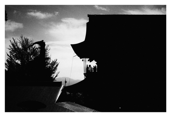 A silhouette of a traditional building against a cloudy sky, with trees in the foreground and mountains in the background. Shadows create a striking contrast in black and white.