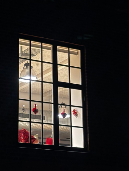 Outside in the dark, looking up at a lit up window in an old factory building, now home to a government agency. Within the panes that make up the tall frame, one can make out the red and white pleated paper of two Danish Christmas hearts. On the wall to the right, a snowflake decoration (there’s also one pasted up against one of the window panes), and on the window sill, what looks like red wrapping paper, a cactus, and a red shopping bag.